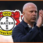 Arne Slot claps his hands with Bayer Leverkusen club crest behind him.