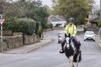 A woman in England riding a paint horse on the road with cars.
