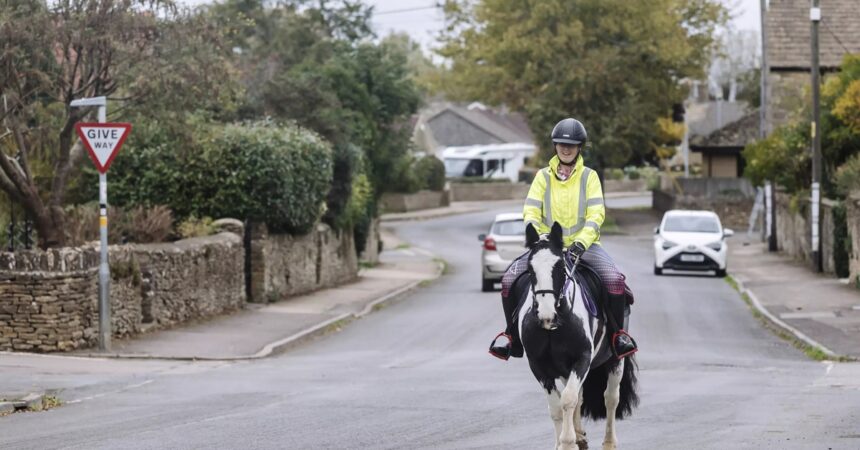 A woman in England riding a paint horse on the road with cars.