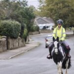 A woman in England riding a paint horse on the road with cars.
