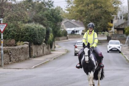 A woman in England riding a paint horse on the road with cars.